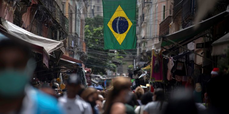 La gente camina en el mercado de Saara bajo una bandera brasileña en el centro de Río de Janeiro, Brasil, el 8 de diciembre de 2020, en medio de la pandemia del coronavirus (COVID-19). - Brasil, el país más poblado de América Latina, tenía 203,1 millones de habitantes en 2022, unos 10 millones menos de lo estimado, según datos del último censo difundido el 28 de junio de 2023 por el instituto oficial de estadística (IBGE). En el estudio demográfico realizado el año pasado se contabilizaron un total de 203.062.512 habitantes. (Foto por Mauro PIMENTEL / AFP)