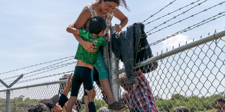 Una madre ayuda a su hijo a cruzar la valla de alambre de púas en Eagle Pass, Texas, después de cruzar a Estados Unidos desde México el 25 de agosto de 2023. (Foto de SUZANNE CORDEIRO / AFP)
